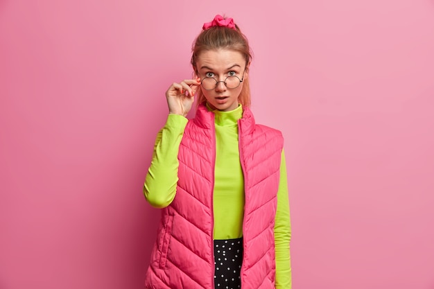 Portrait of shocked adult European woman looks through optical glasses, has astonished expression, keeps hand on rim of spectacles, surprised by what she saw, wears bright clothing