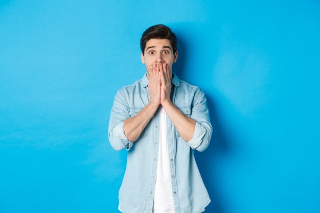 Portrait of shocked adult man looking at camera in awe, gasping and covering mouth with hands, standing over blue background.