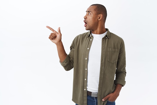 Free photo portrait of shocked alarmed africanamerican handsome guy looking worried and frustrated turn head and pointing finger left gasping scared panic seeing something astounding white background