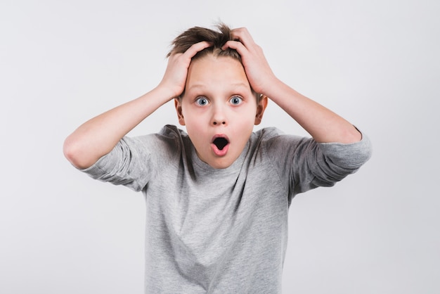 Portrait of a shocked boy with his hands on head looking to camera against grey background