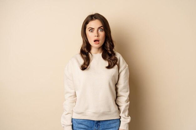 Portrait of shocked brunette woman drop jaw gasping and staring speechless at camera beige background