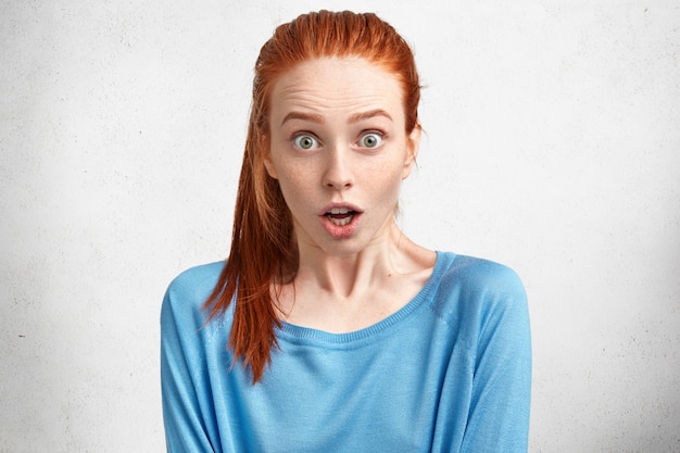 Portrait of shocked bugged eyed pretty redhead young woman expresses great surprisment, hears unexpected news, doesn`t expect to meet somebody