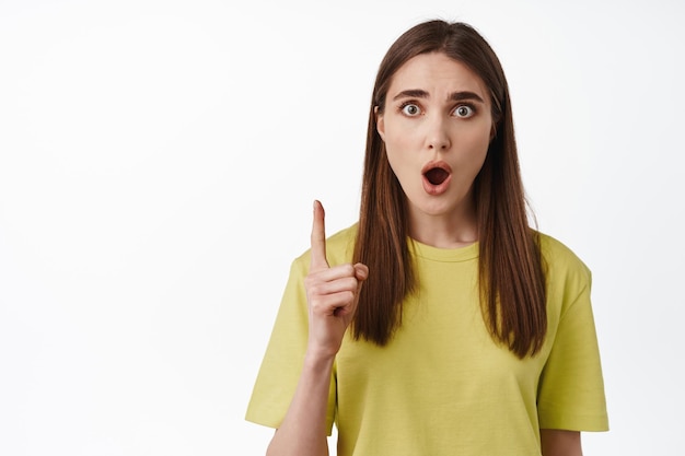 Portrait of shocked girl gasp, pointing finger up and staring in awe, impressed by smth, look with disbelief, standing in t-shirt against white background