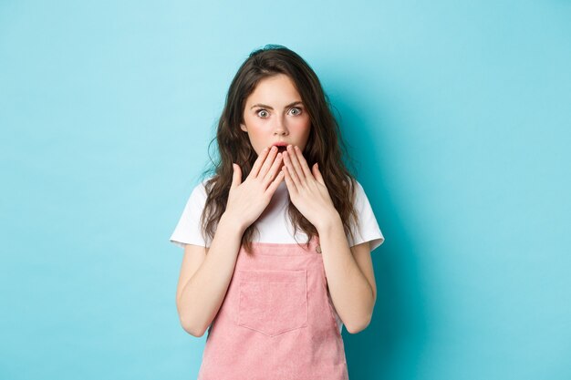 Portrait of shocked gossip girl gasping, covering opened mouth with hands and stare startled at camera, hear rumor, looking with disbelief, standing against blue background.