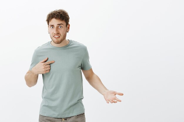 Portrait of shocked offended guy with bristle, gesturing hand and pointing at himself, lifting eyebrow with disbelief