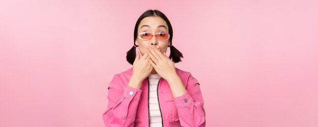 Portrait of shocked stylish asian girl in sunglasses closes mouth looks with surprised face expression stands over pink background Copy space