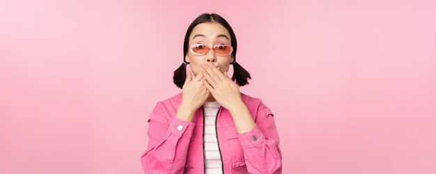 Портрет шокированной стильной азиатской девушки в солнцезащитных очках закрывает рот, смотрит с удивленным выражением лица на розовом фоне.