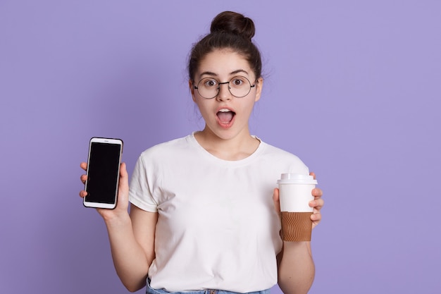 Portrait of shocked surprised woman with dark hair, holding take away coffee cup and mobile phone