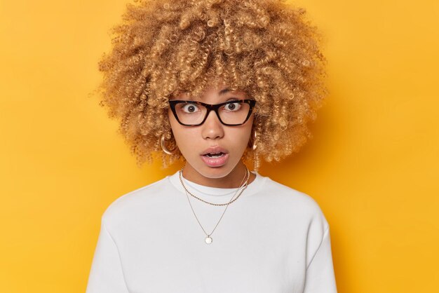 Portrait of shocked woman holds breath and stares wondered at camera wears transparent eyeglasses and white jumper reacts on something amazing isolated over yellow background Human reaction