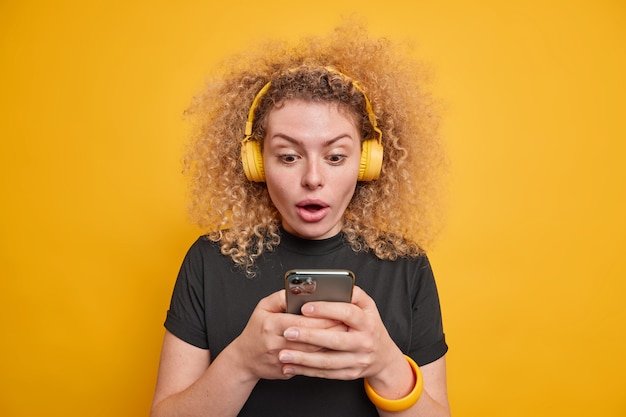 Portrait of shocked woman opens mouth widely focused at smartphone display received unexpected message wears wireless headphones on ears