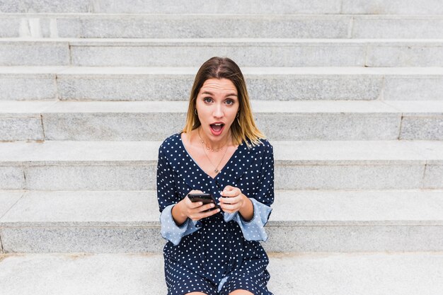 Портрет шокированной женщины, сидя на лестнице, держа мобильный телефон