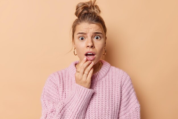 Portrait of shocked young European woman with combed hair keeps mouth opened reacts on shocking news wears casual knitted sweater feels embarrassed poses against brown background Omg concept