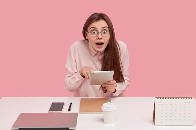 Portrait of shocked young woman does remote work, holds touchpad, makes research for project, keeps jaw dropped