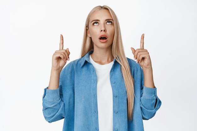 Portrait of shocked young woman drop jaw pointing fingers and looking up with impressed face expression standing over white background