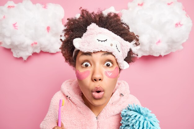 Portrait of shocked young woman has eyes popped out from wonder applies collagen patches under eyes for reducing fine lines poses with toothbrush and bath sponge models indoor