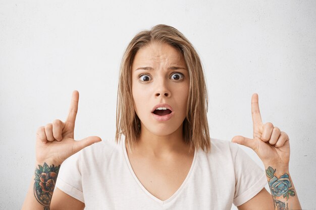 Portrait of shocked young woman with tattoos on arms wearing casual white t-shirt pointing fingers up, having surprised expression, raising eyebrows and opening mouth widely. Horizontal shot