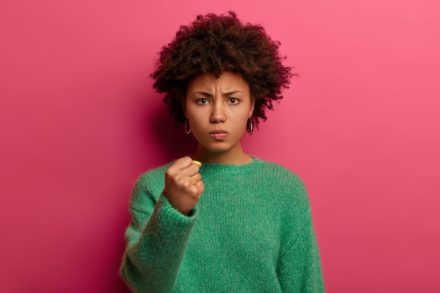 Portrait of short haired angry woman shows fist, has irritated expression, promises to revenge, wears green sweater, poses against pink wall, domineers and threatens, being dissatisfied