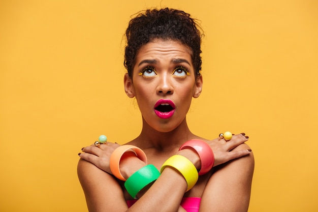 Free photo portrait of shy african woman in colorful adornment with open mouth looking upward crossing hands on shoulders, over yellow wall