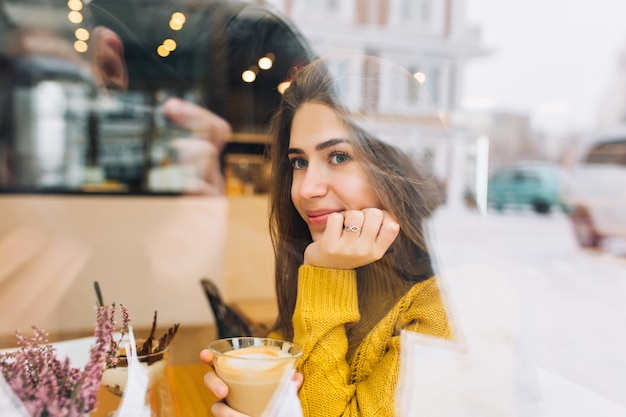 Portrait of shy pensive woman in knitted sweater enjoying coffee and looking at street. Indoor photo of romantic young woman in yellow attire dreaming about something during lunch in cafe.