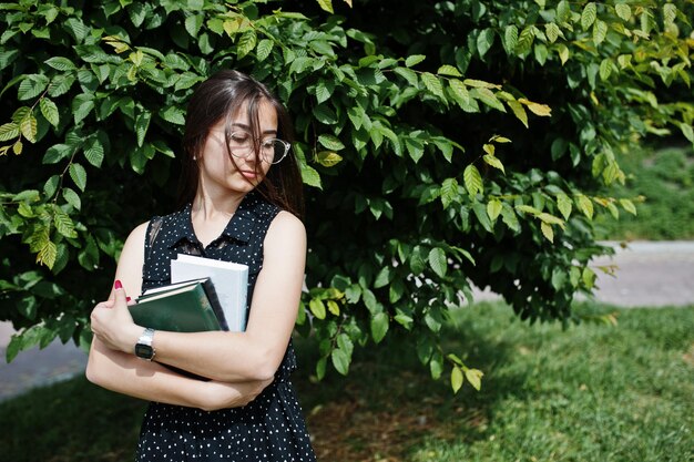 Portrait of a shy young woman in black polka dot dress and glasses holding books in the park