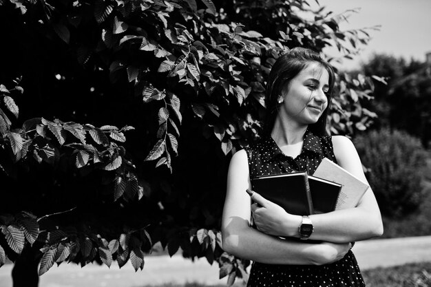 Portrait of a shy young woman in black polka dot dress holding books in the park Black and white photo