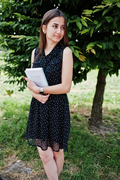 Portrait of a shy young woman in black polka dot dress holding books in the park