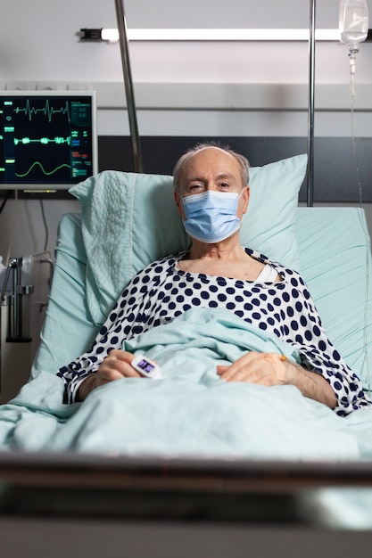 손가락에 산소 농도계를 가진 손에 부착 된 IV 물방울과 함께 병원 침대에서 쉬고 chirurgical 마스크와 아픈 수석 남자 환자의 초상화