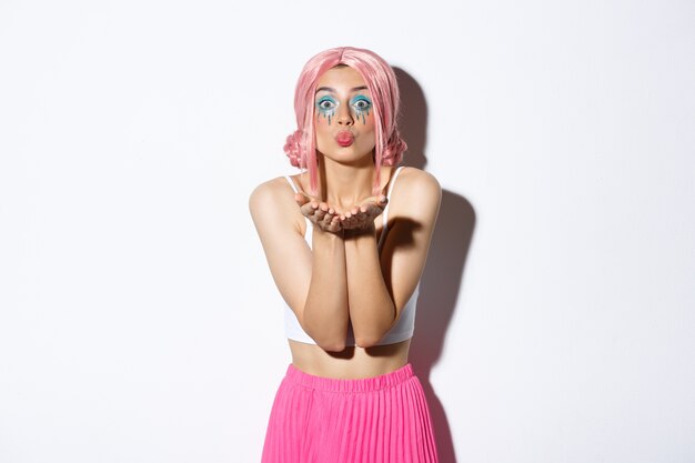 Portrait of silly beautiful caucasian girl with bright makeup, dressed for party in glam pink wig, blowing air kiss at camera, standing.