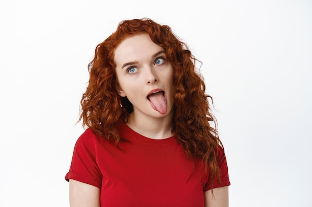 Free photo portrait of silly ginger girl showing tongue and fool around, making funny faces and staring aside, standing against white wall