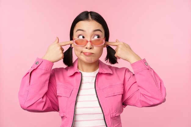 Free photo portrait of silly japanese girl shuts ears looks aside doesnt listen cant hear you gesture standing over pink background ignoring you