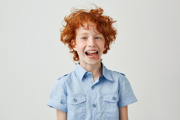 Portrait of silly little ginger boy in blue shirt with wild hair mowing eyes, smiling and showing tongue, making funny faces.