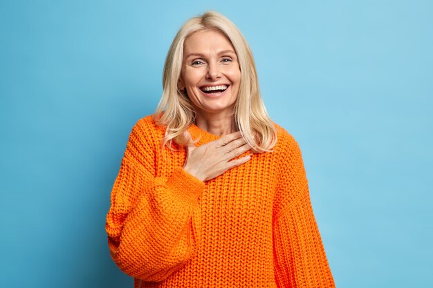 Portrait of sincere blonde woman smiles broadly has white perfect teeth keeps hand on chest feels very glad to receive help from close person wears orange knitted sweater.