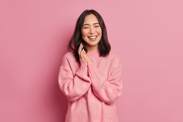 Portrait of sincere brunette Asian female model rubs palms and smiles broadly expresses happiness feels joyful has white even teeth wears oversized casual sweater 