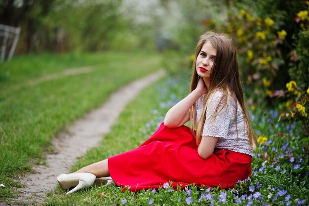 빨간 드레스와 흰 블라우스에 꽃을 입고 풀밭에 있는 봄꽃 정원에서 빨간 입술을 가진 아름다운 소녀의 초상화