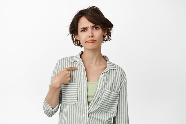 Portrait of skeptical brunette woman smirk, frowning doubtful, pointing at herself with upset, confused face, standing on white.