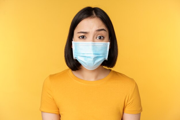 Portrait of skeptical and confused asian woman in medical face mask raising eyebrow doubtful standing over yellow background