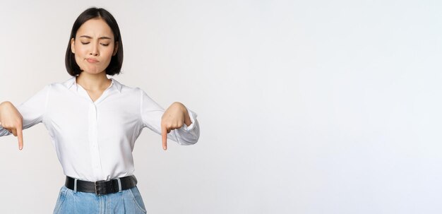 Portrait of skeptical disappointed asian businesswoman korean girl pointing and looking down with doubts standing against white background