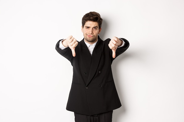 Portrait of skeptical and disappointed man in black suit, frowning upset, showing thumbs-down, dislike something bad, standing over white background.