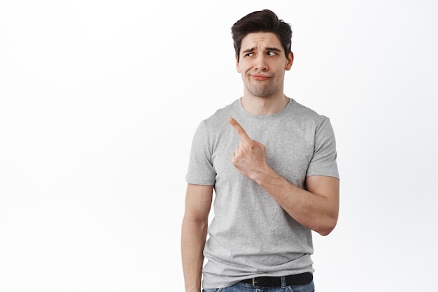 Portrait of skeptical and doubtful guy pointing left and looking with dislike dont like something standing over white background