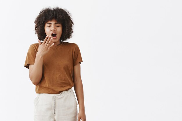 Portrait of sleepy cute African American woman with afro hairstyle closing eyes and yawning with wide opened mouth covering it with hand wanting sleep being tired over gray wall