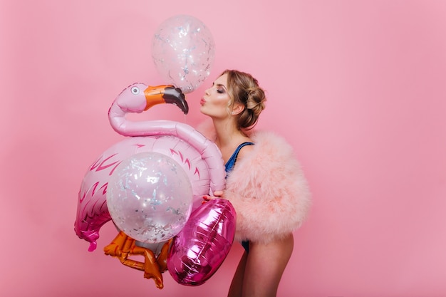 Portrait of slim graceful girl with short blonde hair kissing pink inflatable flamingo. Gorgeous cheerful young woman in fluffy coat having fun with sparkle balloons, waiting for party