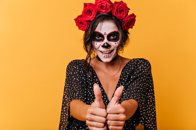 Portrait of sly brown-eyed brunette showing thumbs up. Girl with crown of roses and face art in form of skull smiling.