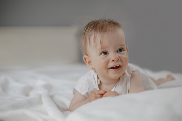 Portrait of smiled wondered little cute baby girl with big eyes laying on the white bedsheet.