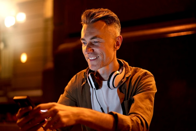 Free photo portrait of smiley man in the city at night with headphones and smartphone