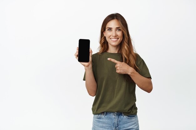 Портрет улыбающейся взрослой женщины, указывающей пальцем на экран мобильного телефона, показывающей интерфейс, рекомендуемое приложение, веб-сайт магазина или компании, новую функцию в приложении, белый фон.