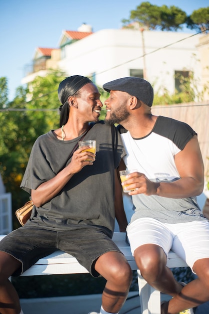 Портрет улыбающейся африканской гей-пары, сидящей на скамейке. Двое мужчин в футболках разговаривают, держа стаканы сока и глядя друг другу в губы для поцелуя. Свободные отношения и любовь к концепции ЛГБТ-пар