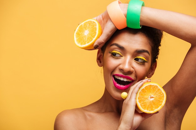 Portrait of smiling afro american woman with stylish makeup holding two halves of juicy orange in both hands isolated, over yellow wall