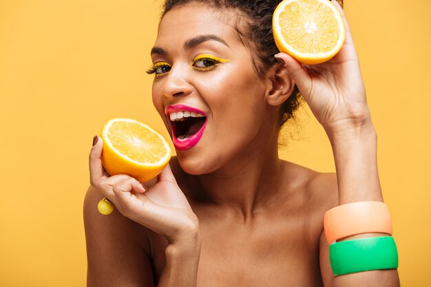 Portrait of smiling afro american woman with stylish makeup tasting juicy ripe orange holding parts in both hands near face isolated, over yellow wall