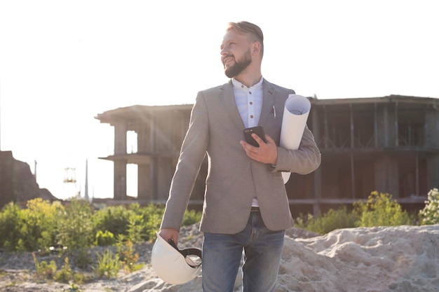 Portrait of smiling architect man holding cellphone and hard hat with blueprint at construction site