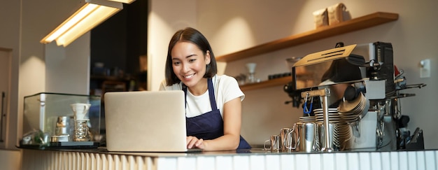 Portrait of smiling asian barista cafe owner entrepreneur working on laptop processing orders on